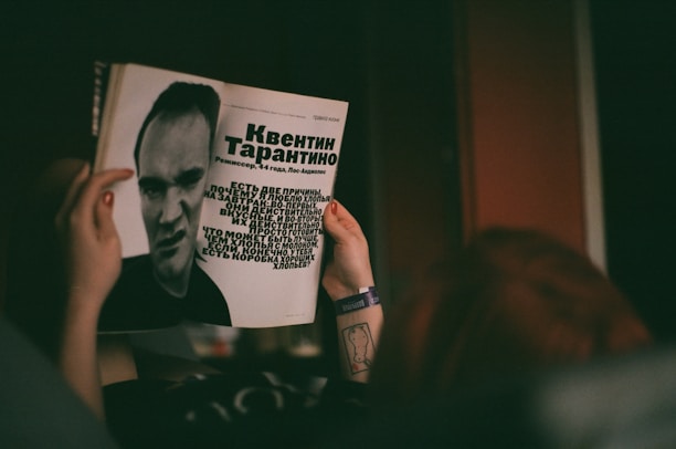 A person is holding a magazine featuring a black-and-white photograph and text about a man. The layout includes large text in a bold font and a stylized portrait of the individual. The person's hands are visible, with a tattoo on the wrist and a wristband.