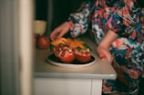 A person wearing a colorful floral-patterned outfit is standing near a table, holding what appears to be a sliced pomegranate on a plate. Additional fruits, including whole pomegranates and slices of an orange-yellow fruit, are arranged on the table next to a white cup.