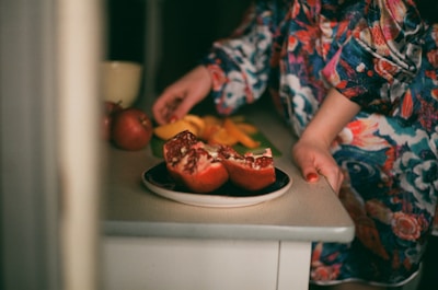 A person wearing a colorful floral-patterned outfit is standing near a table, holding what appears to be a sliced pomegranate on a plate. Additional fruits, including whole pomegranates and slices of an orange-yellow fruit, are arranged on the table next to a white cup.