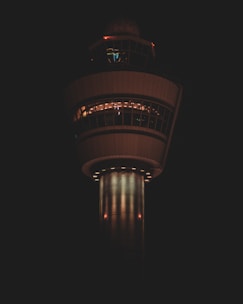 A tall control tower is illuminated against a dark background. The structure has a cylindrical base and a wider observation deck with windows encircling the top. Lights are visible through the windows, creating a warm glow.