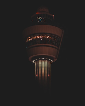 A tall control tower is illuminated against a dark background. The structure has a cylindrical base and a wider observation deck with windows encircling the top. Lights are visible through the windows, creating a warm glow.
