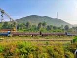 A train carrying stacks of concrete sleepers is situated on tracks amidst lush greenery and hills in the background. Overhead electric lines and structures are visible, indicating an electrified railway line. The area is surrounded by dense foliage and palm trees under a clear blue sky.