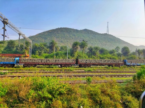 A train carrying stacks of concrete sleepers is situated on tracks amidst lush greenery and hills in the background. Overhead electric lines and structures are visible, indicating an electrified railway line. The area is surrounded by dense foliage and palm trees under a clear blue sky.