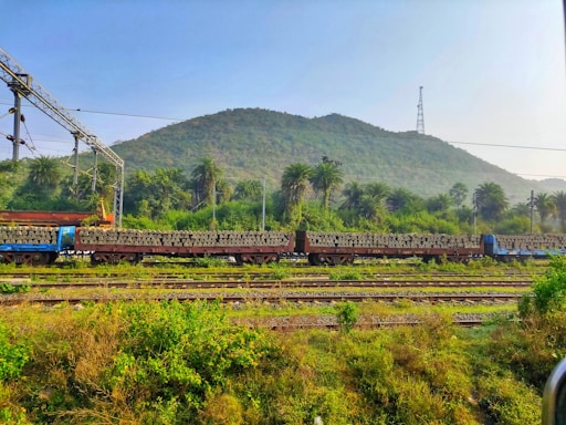 A train carrying stacks of concrete sleepers is situated on tracks amidst lush greenery and hills in the background. Overhead electric lines and structures are visible, indicating an electrified railway line. The area is surrounded by dense foliage and palm trees under a clear blue sky.