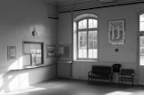 A small waiting area with minimal furnishings is depicted. There is a ticket or information window with a counter, several posters on the walls, and a pair of chairs alongside a radiator under a large window. Sunlight filters through the windows, casting shadows on the floor.