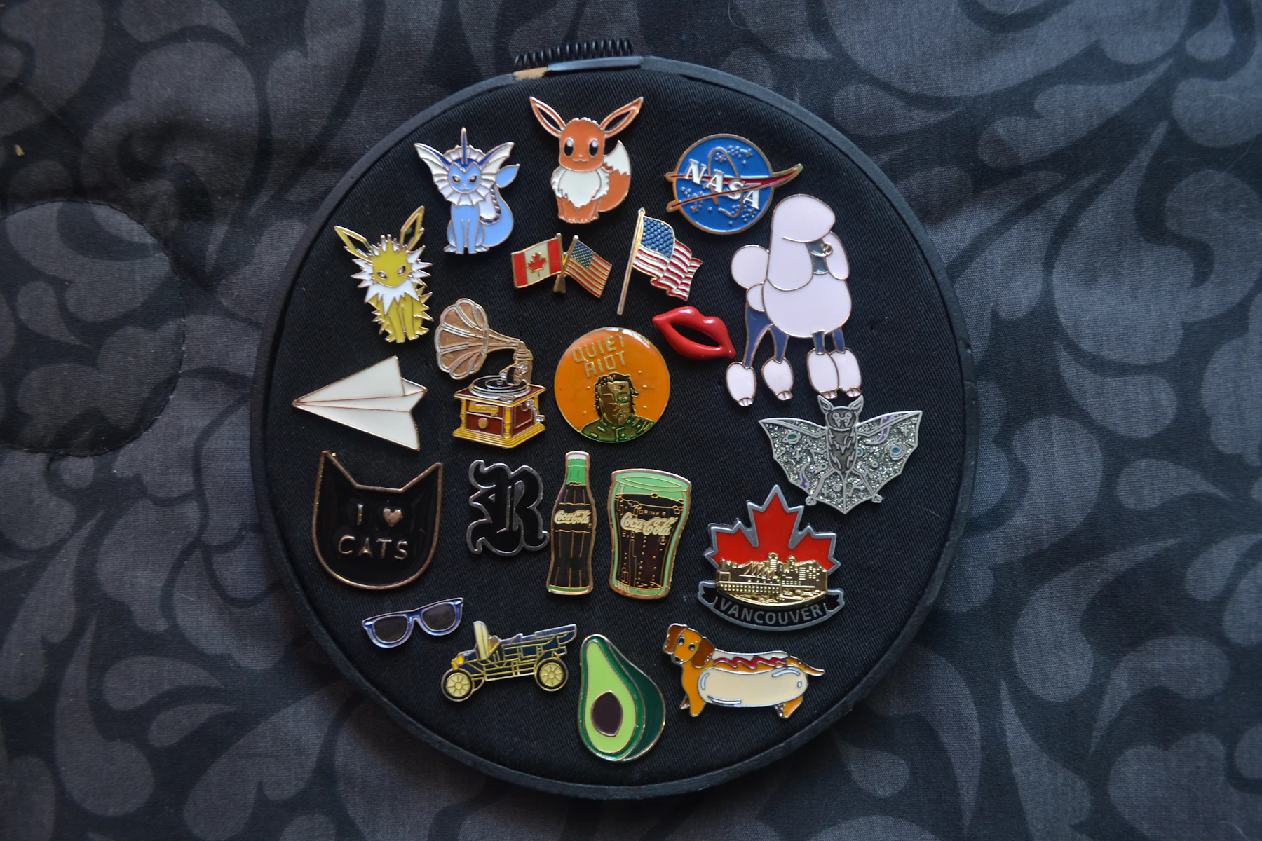 Two dozen enamel pins arranged on black cloth stretched in an embroidery hoop