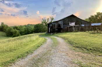 A rustic barn sits atop a small hill surrounded by lush greenery and open fields. An American flag is prominently displayed on the side of the barn. The sky is a mix of soft pastels as the sun sets, casting a warm glow over the landscape. A gravel path winds toward the barn, bordered by well-maintained grass.