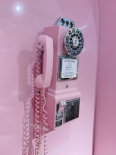 A vintage-style rotary phone mounted on a wall, featuring a pink color scheme with shiny metallic accents. The phone includes a coiled cord and a coin slot for calls.