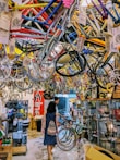 A busy bicycle shop filled with colorful bike frames hanging from the ceiling. Two people walk through the space, exploring the various bike parts and accessories displayed on racks and in glass cases. The overall atmosphere is vibrant and cluttered.