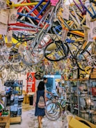 A busy bicycle shop filled with colorful bike frames hanging from the ceiling. Two people walk through the space, exploring the various bike parts and accessories displayed on racks and in glass cases. The overall atmosphere is vibrant and cluttered.