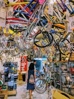 A busy bicycle shop filled with colorful bike frames hanging from the ceiling. Two people walk through the space, exploring the various bike parts and accessories displayed on racks and in glass cases. The overall atmosphere is vibrant and cluttered.