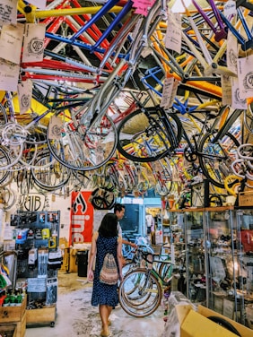 A busy bicycle shop filled with colorful bike frames hanging from the ceiling. Two people walk through the space, exploring the various bike parts and accessories displayed on racks and in glass cases. The overall atmosphere is vibrant and cluttered.