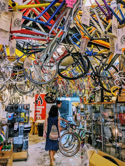 A busy bicycle shop filled with colorful bike frames hanging from the ceiling. Two people walk through the space, exploring the various bike parts and accessories displayed on racks and in glass cases. The overall atmosphere is vibrant and cluttered.