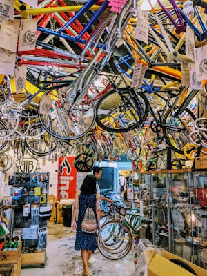 A busy bicycle shop filled with colorful bike frames hanging from the ceiling. Two people walk through the space, exploring the various bike parts and accessories displayed on racks and in glass cases. The overall atmosphere is vibrant and cluttered.