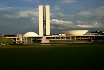 A large and distinctive modernist building complex featuring two prominent symmetrical towers, a dome, and a bowl-shaped structure. The structures are set against a backdrop of a partly cloudy sky, with a vast green lawn in the foreground. The architecture is characterized by clean lines and geometric shapes.