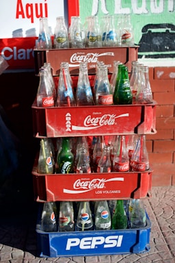 A stack of crates containing empty soda bottles is arranged on a sidewalk. The top three crates are red and branded with Coca-Cola logos, while the bottom crate is blue with a Pepsi logo. Various soda brands such as Pepsi, Coca-Cola, Sprite, and Crush are visible on the bottles.