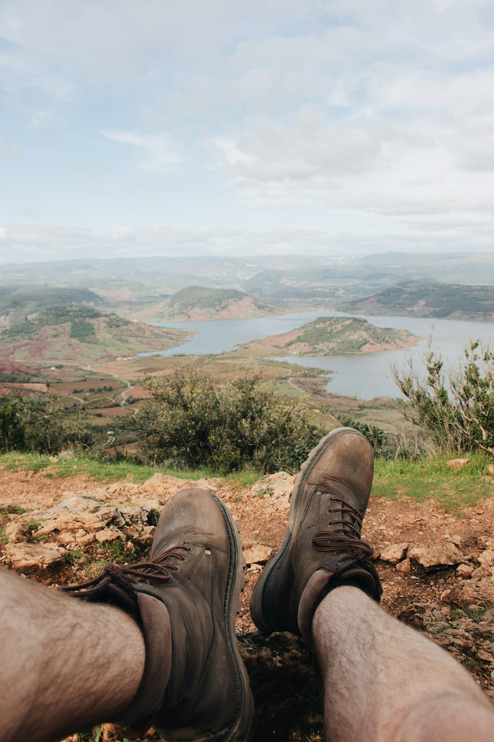 man sitting on rocky mountain facing the mountains and body of water