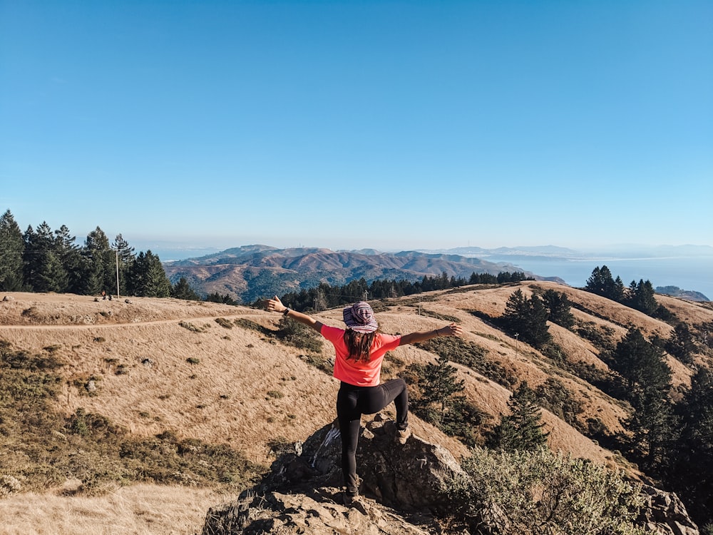 person standing on cliff while raising both hands viewing mountain and body of water under blue and white sky