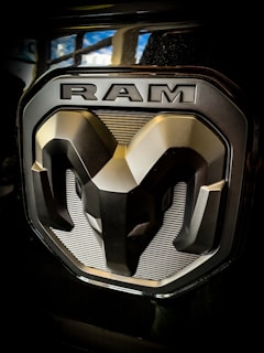 A metallic car emblem featuring a bold, stylized ram head with the word 'RAM' above it. The design is engraved on a shiny, dark background with prominent highlights.