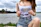 A person outdoors near a body of water, holding sunglasses in both hands. They are wearing a white tank top with a logo that reads 'REAL POKER' in gold letters and denim shorts. The background features a cloudy sky and some trees lining the shore.