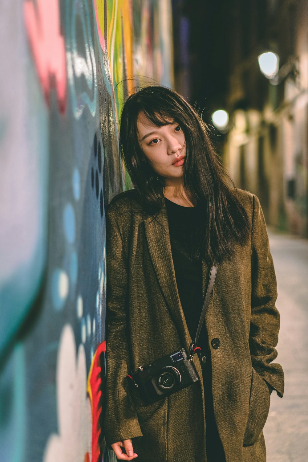 woman wearing brown coat with camera standing and leaning against the wall