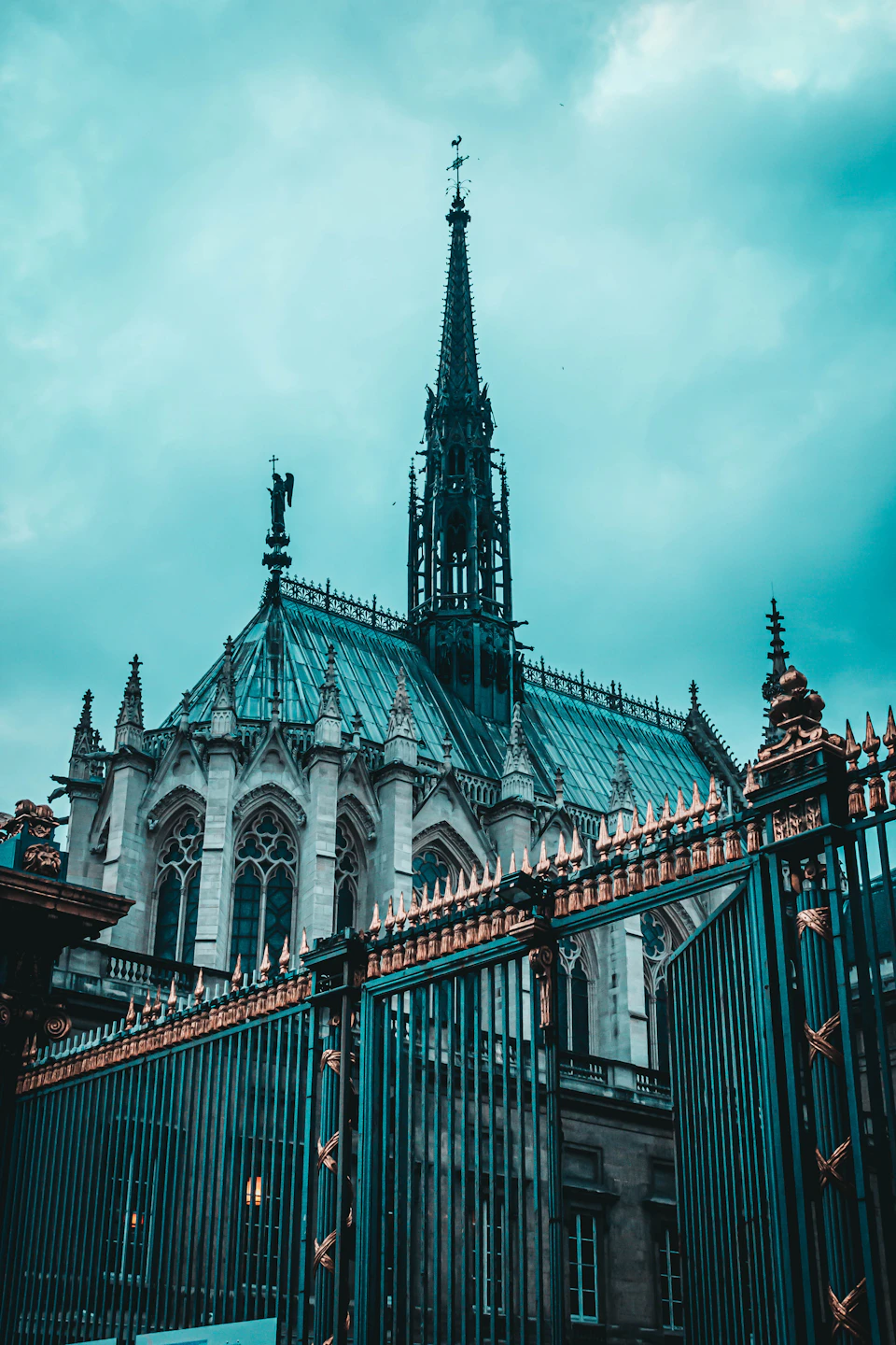 The image depicts a large, ornate church with a tall spire and a statue perched atop it. The church is constructed from stone and features intricate carvings on its facade. A green metal fence surrounds the church, adding an element of modernity to the historic structure. In the foreground, there are trees visible behind the church, providing a natural backdrop to this architectural marvel.