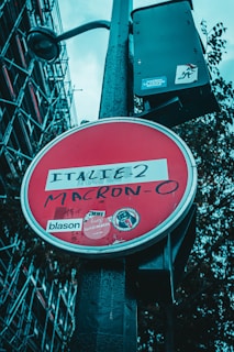 A red circular sign with a white border contains graffiti writing 'ITALIE-2 MACRON-0'. The sign has various stickers, including logos and text, and is mounted on a black pole. In the background, there is a structure with metal scaffolding, and the sky is overcast.