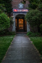 A dimly lit pathway leads to a brick building with an illuminated neon sign that reads 'The Rasmussen' above the entrance. The greenery on either side of the walkway creates a secluded and intimate atmosphere.