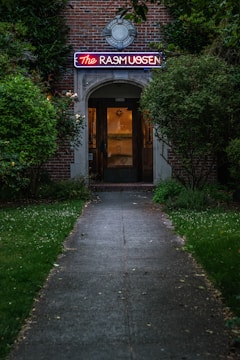 A dimly lit pathway leads to a brick building with an illuminated neon sign that reads 'The Rasmussen' above the entrance. The greenery on either side of the walkway creates a secluded and intimate atmosphere.