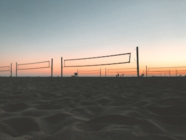 Several beach volleyball nets are set up on a sandy beach during sunset. The sky is a gradient of blue and orange hues, with silhouettes of a few people walking near the nets.