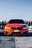 A bright orange sports car with a prominent front grille is parked on a road. The car has a license plate reading 'LDZ 191'. In the background, various people are gathered, some standing near other parked vehicles under a clear blue sky.