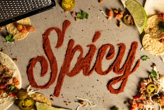 The word 'Spicy' is written in red chili powder on a gray surface, surrounded by tortilla chips, lime wedges, sliced jalapeños, grated cheese, cilantro, and chopped tomatoes, evoking a vibrant culinary theme.