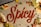 The word 'Spicy' is written in red chili powder on a gray surface, surrounded by tortilla chips, lime wedges, sliced jalapeños, grated cheese, cilantro, and chopped tomatoes, evoking a vibrant culinary theme.