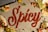 The word 'Spicy' is written in red chili powder on a gray surface, surrounded by tortilla chips, lime wedges, sliced jalapeños, grated cheese, cilantro, and chopped tomatoes, evoking a vibrant culinary theme.