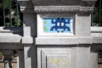 A stone wall features a small mosaic tile artwork. The mosaic resembles a pixelated character, reminiscent of vintage video games, in shades of blue and white. The weathered stone frame has decorative carvings, and some graffiti is visible at the bottom in yellow.