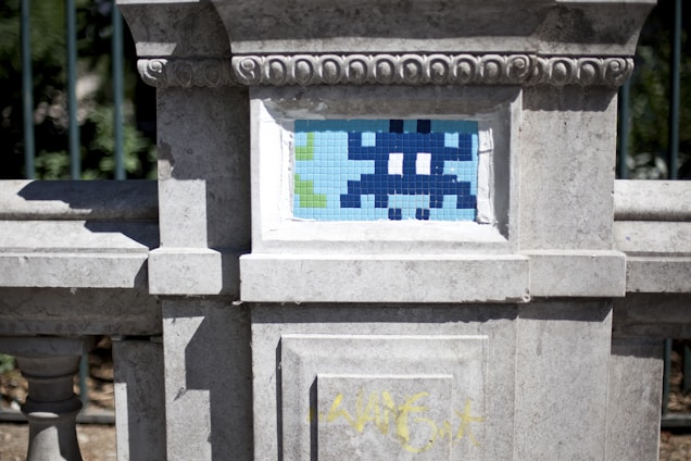 A stone wall features a small mosaic tile artwork. The mosaic resembles a pixelated character, reminiscent of vintage video games, in shades of blue and white. The weathered stone frame has decorative carvings, and some graffiti is visible at the bottom in yellow.