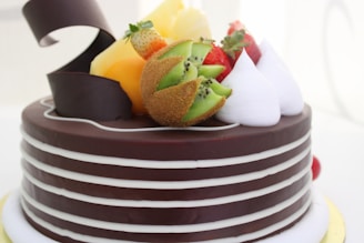 A chocolate cake decorated with layers of white stripes and topped with a variety of fresh fruits including strawberries, kiwi slices, and pineapple. Whipped cream peaks are also present along with a decorative chocolate curl.