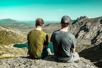 two men sitting on cliff during daytime