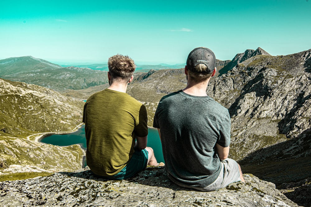 two men sitting on cliff during daytime