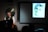 A person sits in front of a laptop, with a large gong behind them, delivering a presentation. The room is dimly lit, and a black-and-white portrait of a woman is projected onto a screen beside them. The audience is visible in silhouette.