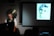 A person sits in front of a laptop, with a large gong behind them, delivering a presentation. The room is dimly lit, and a black-and-white portrait of a woman is projected onto a screen beside them. The audience is visible in silhouette.