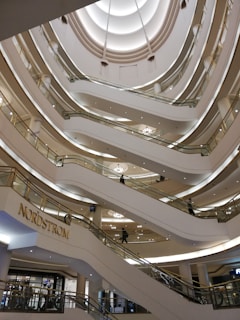Multiple levels of an indoor shopping mall with modern architectural design. Numerous escalators connect the floors, and a store sign for Nordstrom is prominently displayed. The ceiling features an intricate pattern with soft lighting.