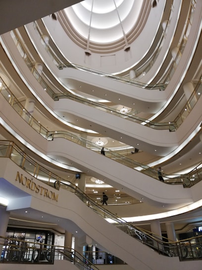 Multiple levels of an indoor shopping mall with modern architectural design. Numerous escalators connect the floors, and a store sign for Nordstrom is prominently displayed. The ceiling features an intricate pattern with soft lighting.
