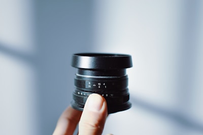A person is holding a black camera lens with visible aperture and focus markings on it. The background is softly lit and out of focus, creating a minimalist and clean aesthetic.