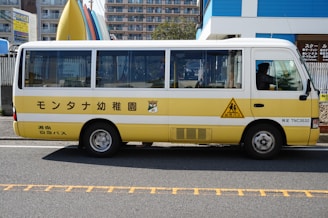 A small, yellow and white bus is parked on the side of a road. The bus has Japanese text written on the side and is likely intended for transporting children, as indicated by the child crossing logo. In the background, there are buildings and a fence, along with colorful surfboards on display.