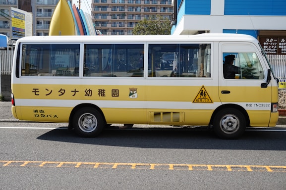 A small, yellow and white bus is parked on the side of a road. The bus has Japanese text written on the side and is likely intended for transporting children, as indicated by the child crossing logo. In the background, there are buildings and a fence, along with colorful surfboards on display.