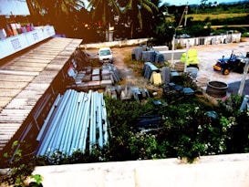 An outdoor storage area with a variety of construction materials, including metal pipes, slabs, and blocks. A small van is parked on the property, and there are large concrete rings lying on the ground. The area is surrounded by greenery and trees, with a building on one side and an open field in the background.