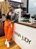 A person wearing an all-black outfit with a denim jacket over a black top, orange cargo pants, and white sneakers. They have accessorized with sunglasses, large hoop earrings, multiple rings, and a headscarf. The individual is leaning casually against a store counter labeled 'NANA JUDY', with shelves displaying sunglasses behind them and various folded clothing items and accessories visible in the background.