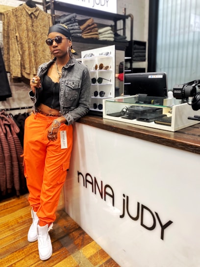 A person wearing an all-black outfit with a denim jacket over a black top, orange cargo pants, and white sneakers. They have accessorized with sunglasses, large hoop earrings, multiple rings, and a headscarf. The individual is leaning casually against a store counter labeled 'NANA JUDY', with shelves displaying sunglasses behind them and various folded clothing items and accessories visible in the background.