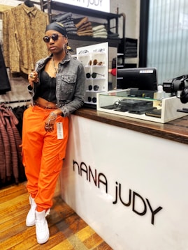 A person wearing an all-black outfit with a denim jacket over a black top, orange cargo pants, and white sneakers. They have accessorized with sunglasses, large hoop earrings, multiple rings, and a headscarf. The individual is leaning casually against a store counter labeled 'NANA JUDY', with shelves displaying sunglasses behind them and various folded clothing items and accessories visible in the background.
